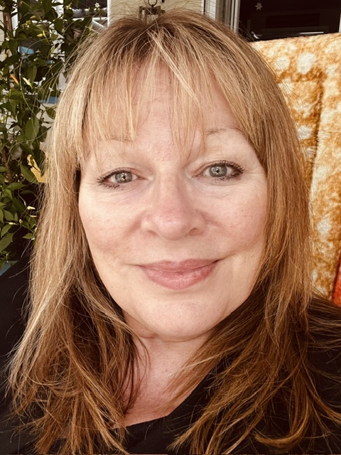Close-up portrait of a person with shoulder-length light brown hair, wearing a black top, with indoor plants and a patterned throw in the background. Close-up portrait of a person with shoulder-length light brown hair, wearing a black top, with indoor plants and a patterned throw in the background.