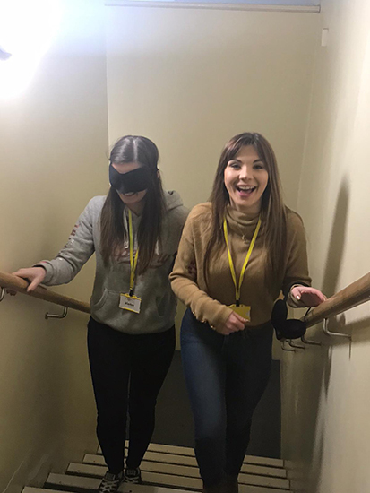 Two individuals going trough a sighted guide training taster, one sighted person leading the other who is blindfolded up a flight of stairs 
