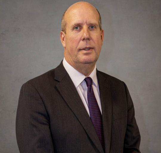 Professional head-and-shoulders portrait of a person wearing a dark business suit, light shirt and patterned tie, standing against a plain grey background Professional head-and-shoulders portrait of a person wearing a dark business suit, light shirt and patterned tie, standing against a plain grey background