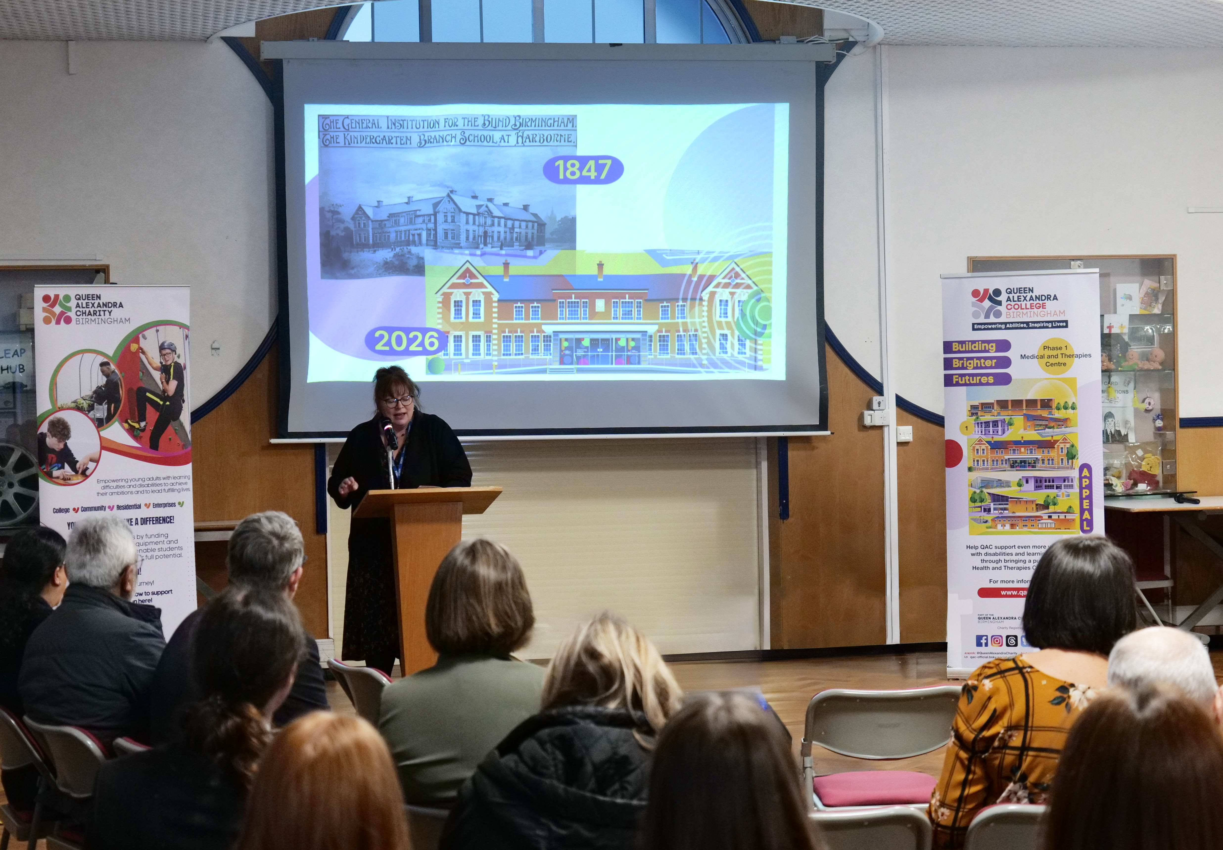 Image of Principal, Bev talking to an audience with a presentation shows in the background