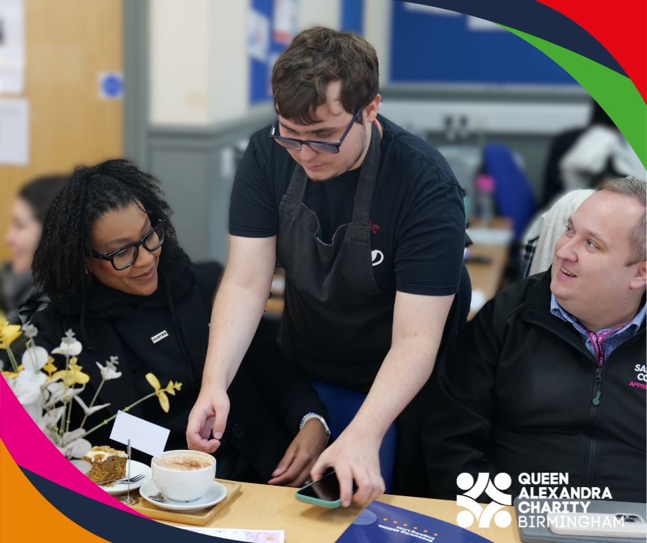 A male student wearing an apron serves a cup of coffee to two event attendees one male one female seated at a café-style table. Flowers, cake, and warm lighting create a welcoming atmosphere. The Queen Alexandra Charity Birmingham logo appears in the corner.