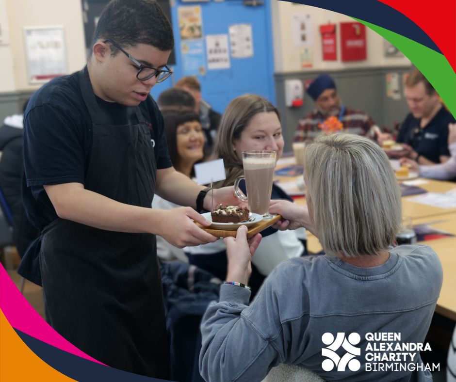 A male hospitality student hands a slice of cake and a tall glass of hot chocolate to a woman event attendee seated at the café table. People in the background. The Queen Alexandra Charity Birmingham logo is in the corner.
