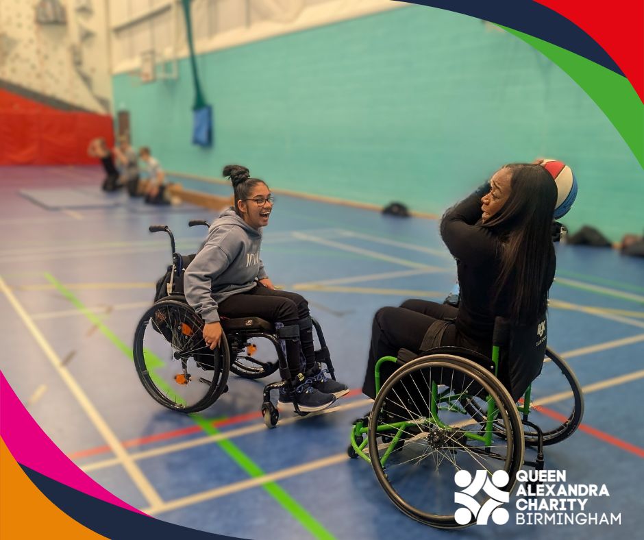 One student and one event attendee playing in sports wheelchairs laugh together while taking part in wheelchair basketball. One holds a blue-and-white ball ready to pass it. The Queen Alexandra Charity Birmingham logo is in the corner.