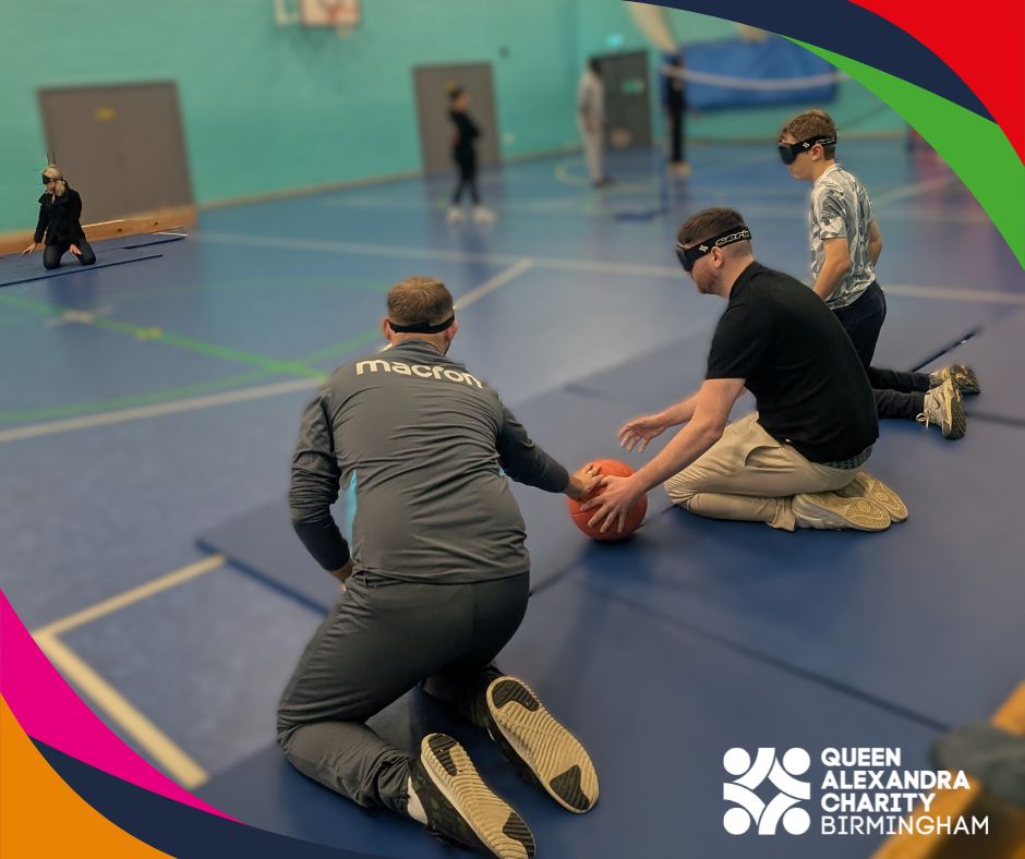 Three blindfolded men, two event attendees one student kneel on a sports hall floor while playing goal ball two catching orange ball. Other participants in the background. Colourful curved borders frame the image, with the Queen Alexandra charity Birmingham logo in the corner.