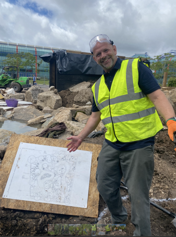 A person wearing a high-visibility yellow vest, gloves, and safety goggles A person wearing a high-visibility yellow vest, gloves, and safety goggles stands on a construction site at the NEC, gesturing toward a garden design plan mounted on a board