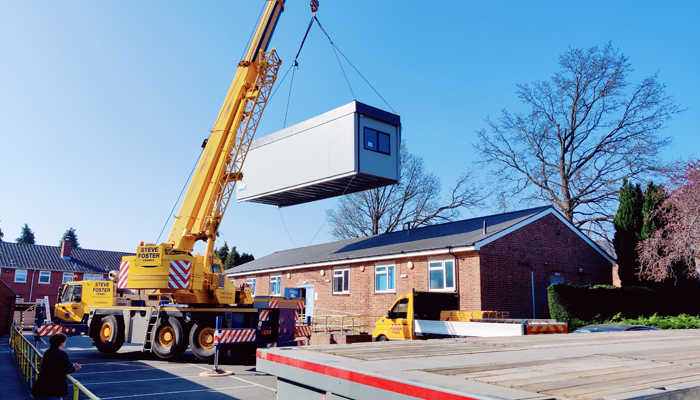 The modular building pictured being hoisted over a building on QAC campus