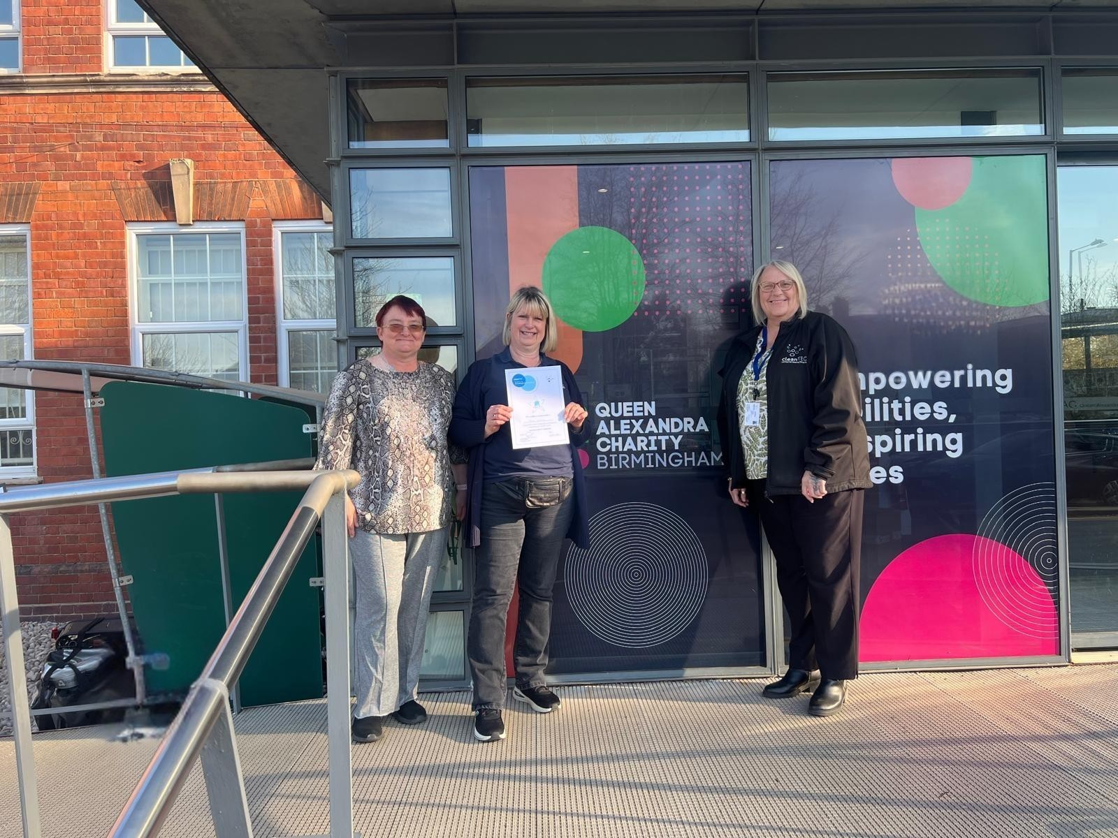 Image of Tina and two members of staff outside the reception of QAC holding a certificate